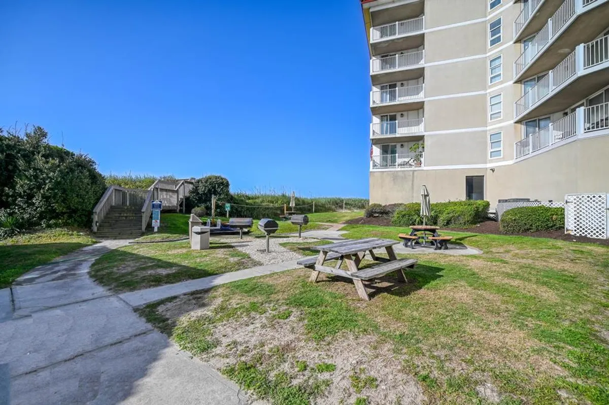 An outdoor grilling and picnic area with picnic tables, a grilling station, and a staircase leading down to the beach. The open-air setting encourages socializing, dining, and relaxation with ocean views.