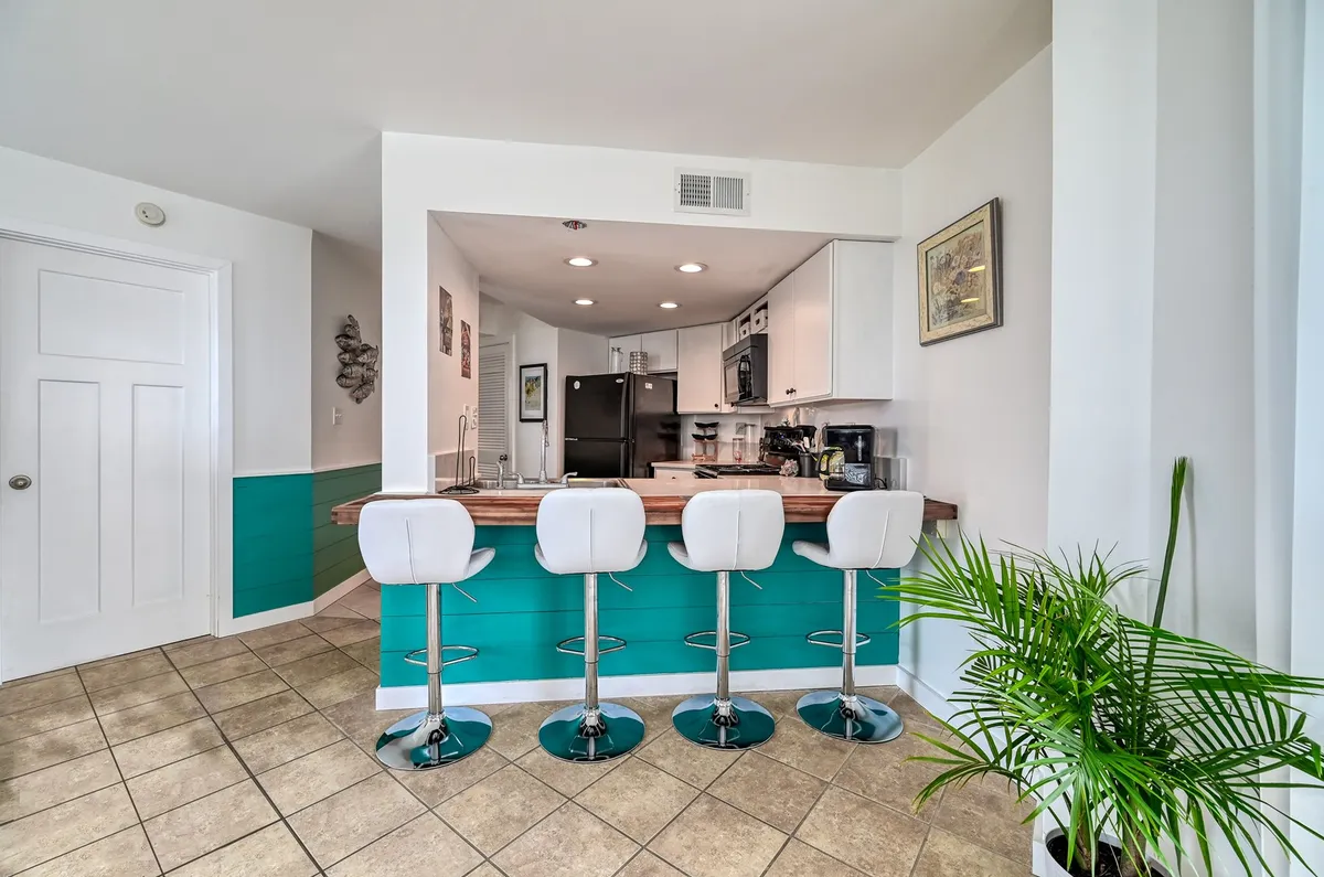 A bright, open kitchen with a central island flanked by bar stools, a green accent wall, and a potted plant adding a modern coastal vibe. The space is designed for cooking, casual dining, and lively social interaction.