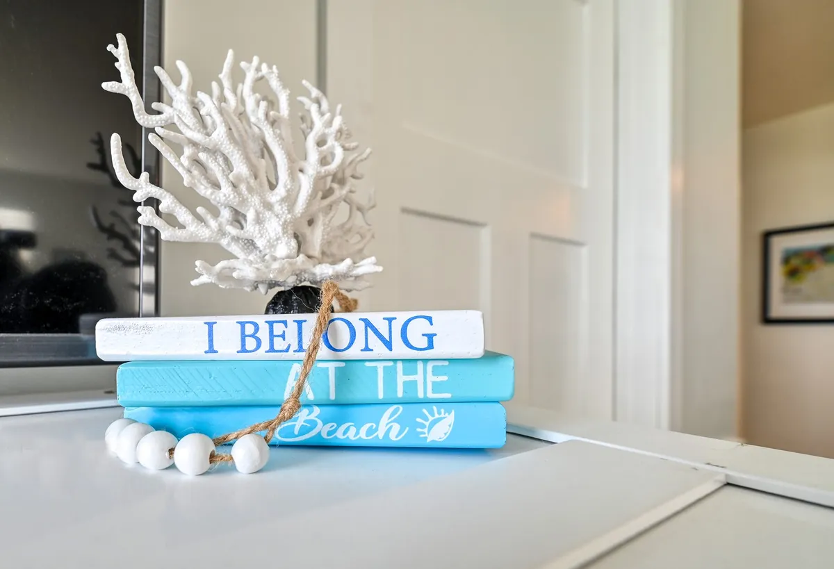 A cozy reading nook featuring a stack of blue books and a white coral decoration atop a beaded table. The decor is coastal-inspired and relaxing, inviting guests to unwind and enjoy a good book.