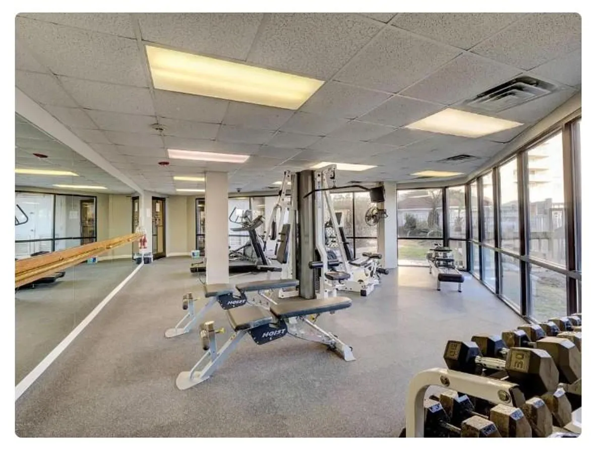 A bright and well-equipped fitness center with weightlifting equipment and exercise machines is showcased. Large windows provide plenty of natural light, giving the gym an airy and motivational atmosphere. The design suggests a modern space ideal for workouts and group fitness activities.