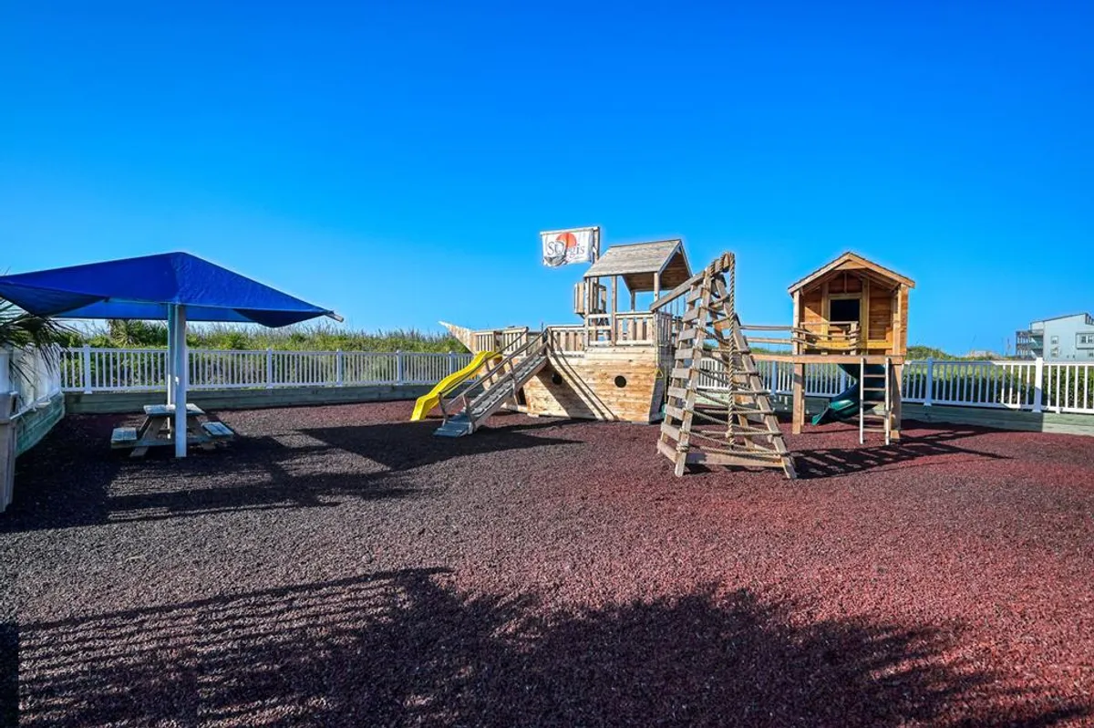A colorful playground structure nestled in a sand area, complete with slides, a climbing frame, and a sunshade. Children play and families gather, creating a joyful, safe environment for imaginative play and outdoor bonding.