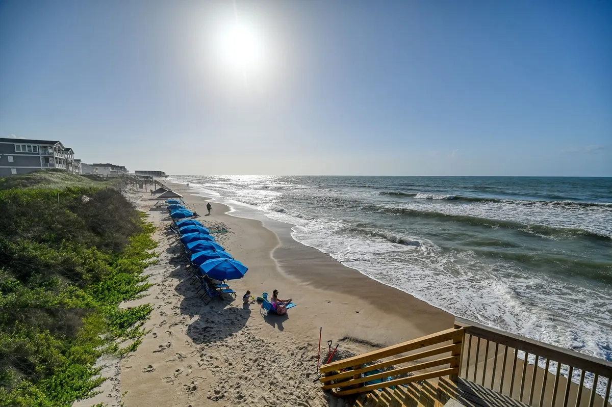 A lively beachfront scene with families and children playing in the sand, colorful beach umbrellas, sparkling ocean waves, and bright sunshine. The energy captures classic beach activities like sandcastle building, swimming, and beach games.