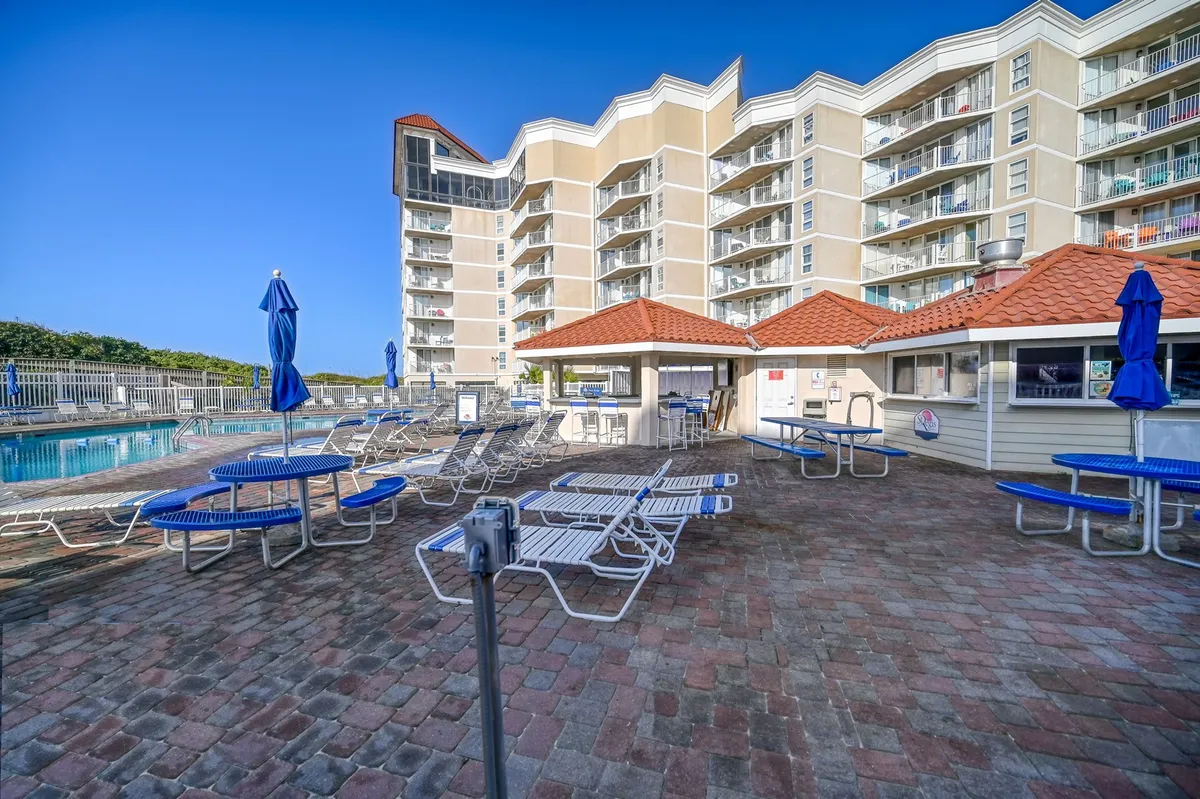 This image features a sparkling swimming pool surrounded by lounge chairs, umbrellas, and a cabana, set against the facade of a resort-style building. The setting radiates a sunny, inviting ambiance ideal for both relaxation and socializing.