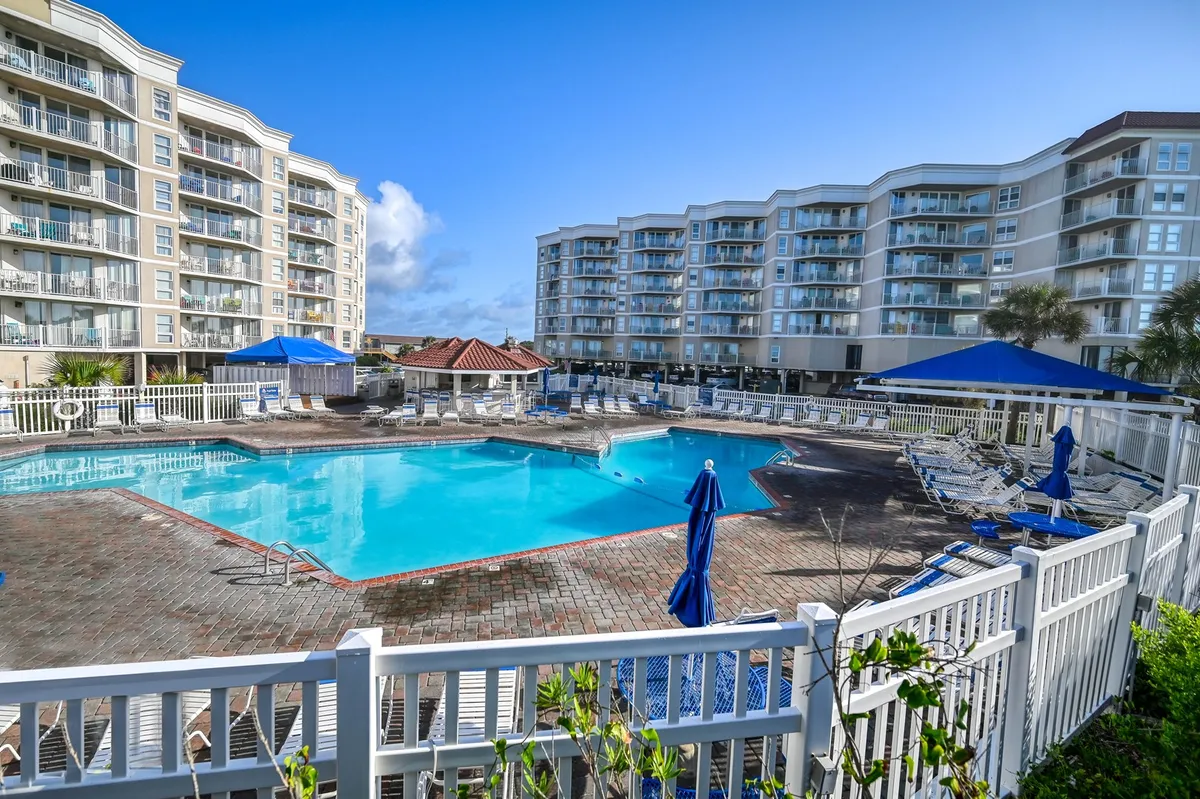 An inviting exterior swimming pool area complete with sun loungers, blue umbrellas, and surrounding resort buildings. The mood is leisurely, with space for both relaxation and socializing, and the pool area is a focal point for gatherings and recreation.