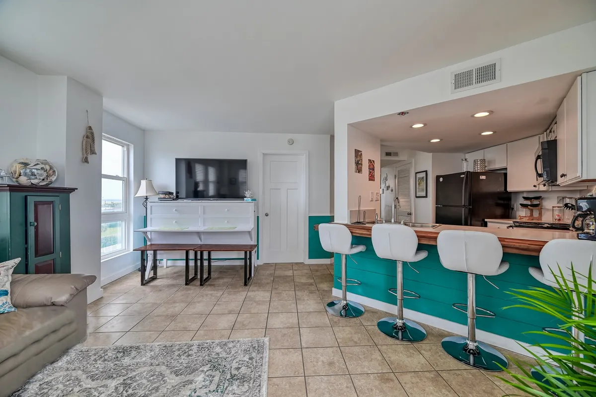 The image showcases a bright, inviting condo interior with a spacious kitchen island and bar stools, ample natural light pouring in through a large window, a cozy TV area, and stylish decorative touches throughout. The scene suggests a space perfect for gathering with friends or family, enjoying home-cooked meals, and relaxing in comfort.