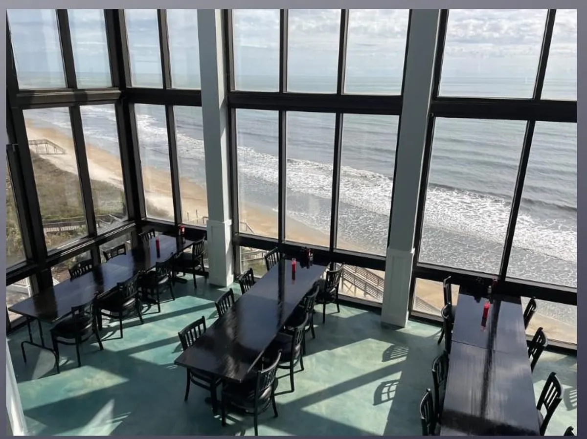 An elegant indoor dining area with large glass windows framing a stunning ocean view. The tables are set for dining, with candles adding ambiance, and the entire scene looks serene and uplifting—perfect for special meals and oceanfront celebrations.