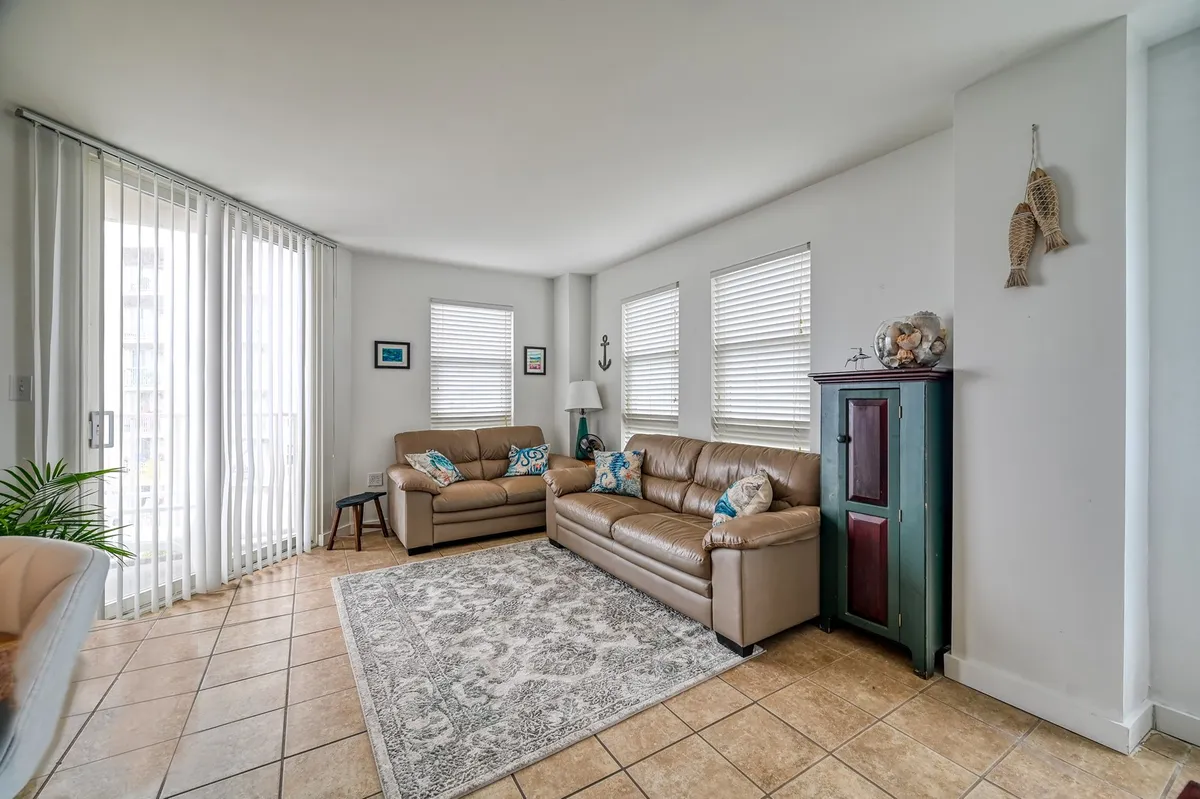 A cozy living room featuring plush leather sofas with decorative cushions, a textured area rug, and abundant natural light streaming through a sliding glass door. A corner cabinet adds a touch of elegance, while the setting feels perfect for relaxed gatherings, socializing, or movie nights.